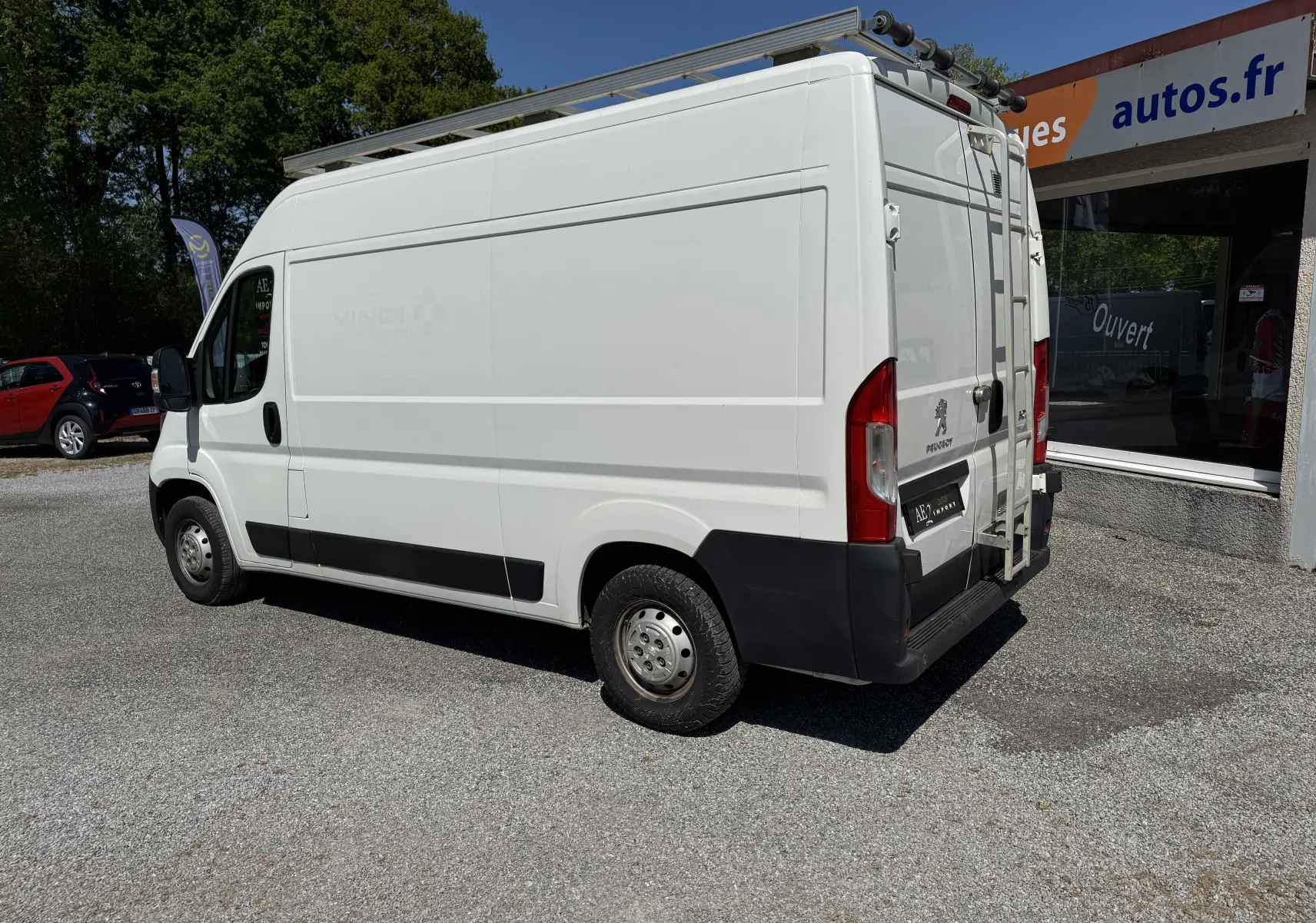Peugeot Boxer Fourgon blanc vu de trois quarts arrière droit avec échelle et galerie sur le toit, stationné devant un garage.