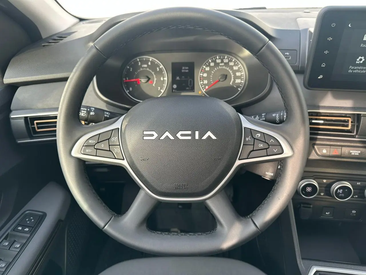 Vue rapprochée du volant noir Dacia avec commandes intégrées, tableau de bord analogique et écran tactile central.