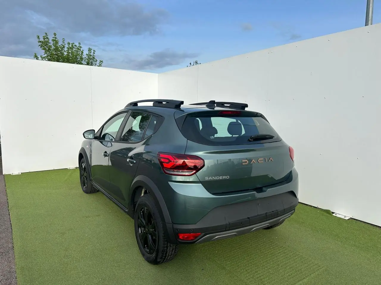 Vue 3/4 arrière droite d'une Dacia Sandero III Stepway 2025 vert cèdre avec barres de toit et jantes noires.