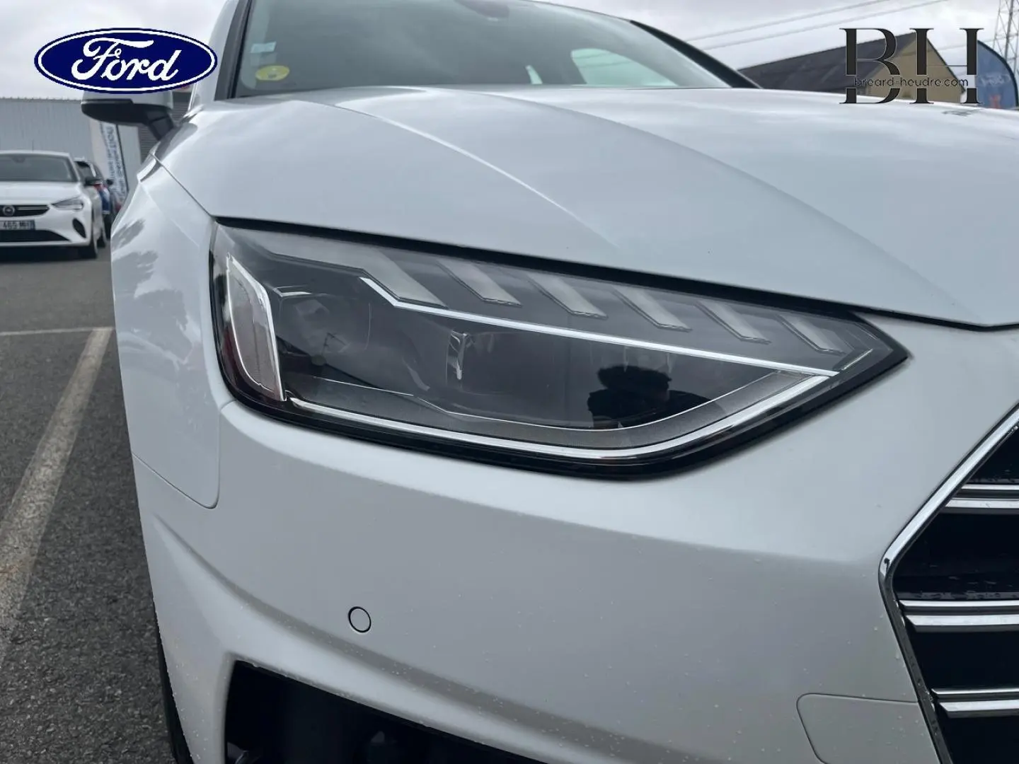 Gros plan sur le phare avant gauche d'une Audi A4 Avant blanc glacier métallisé, avec calandre partielle visible.