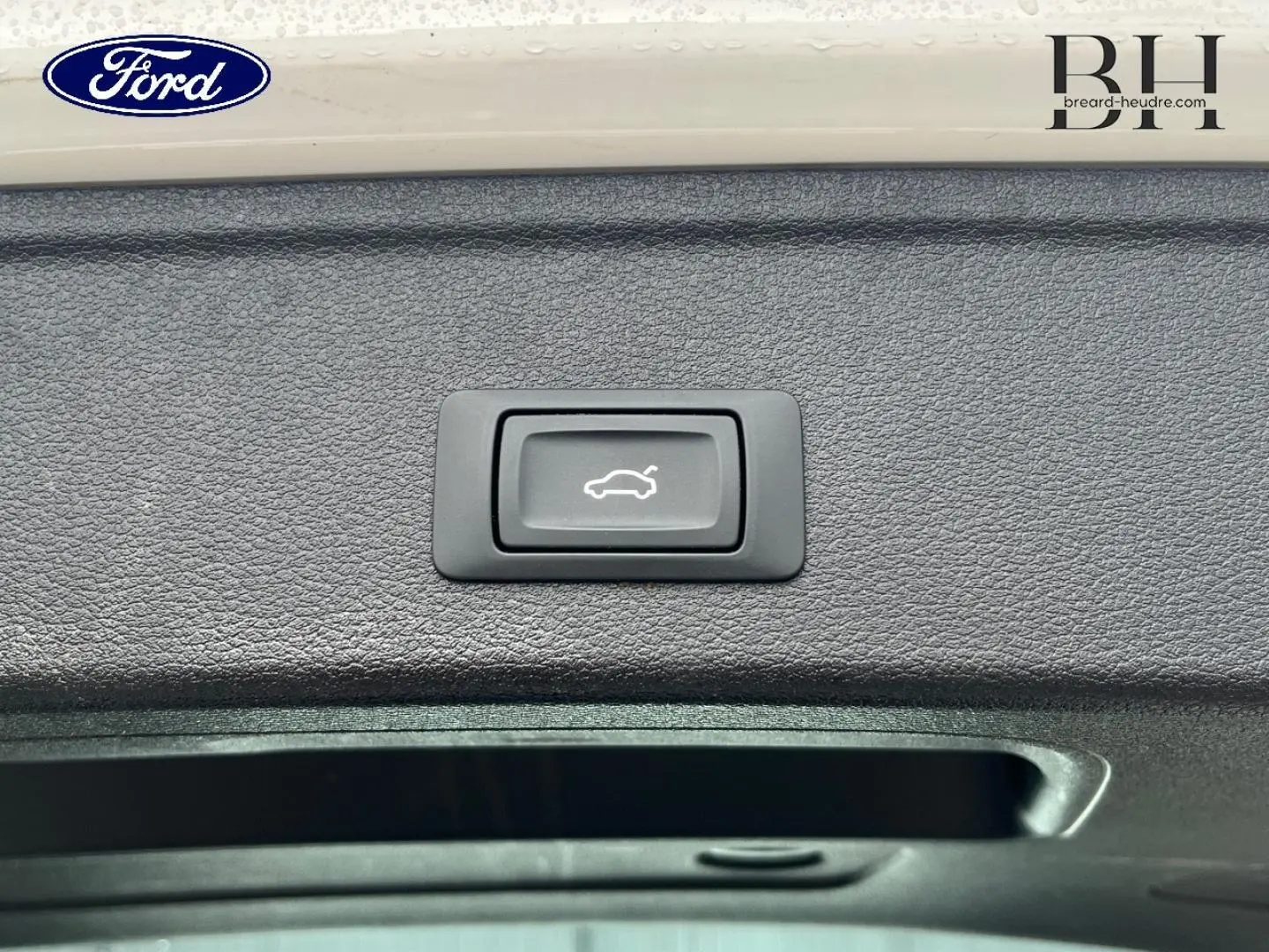 Gros plan sur le bouton d'ouverture électrique du coffre, entouré de garniture noire texturée.