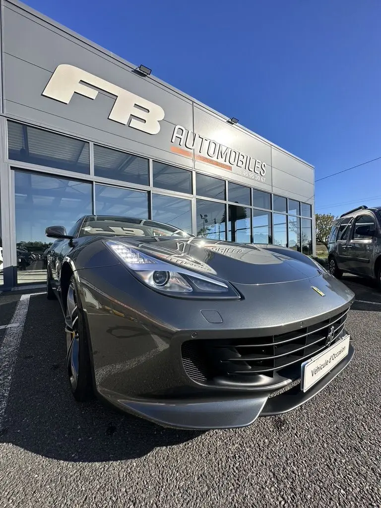 Ferrari GTC4 Lusso gris foncé vue 3/4 avant droit, avec phares allumés devant un showroom FB Automobiles.