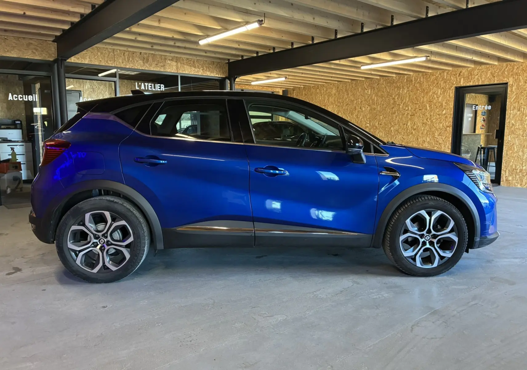 Renault Captur bleu en profil droit dans un garage, avec toit noir et jantes alliage distinctives.