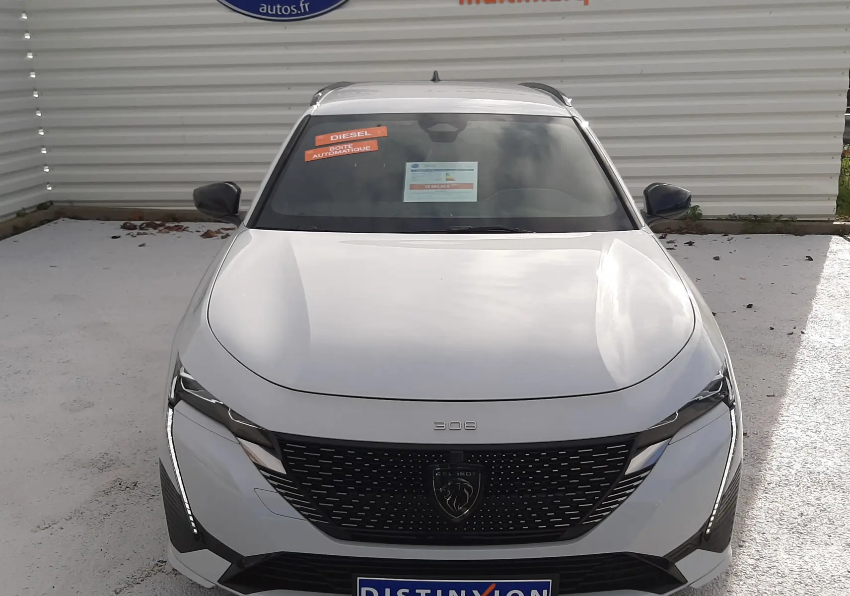 Vue avant d'une Peugeot 308 SW blanche Okenite avec calandre noire et phares LED, stationnée devant un mur publicitaire.