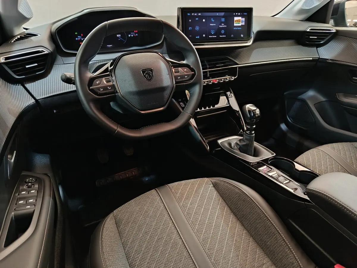 Intérieur de la Peugeot 208 PureTech 100 S&S Allure 2025, vue côté conducteur, volant cuir et console centrale avec boîte manuelle.