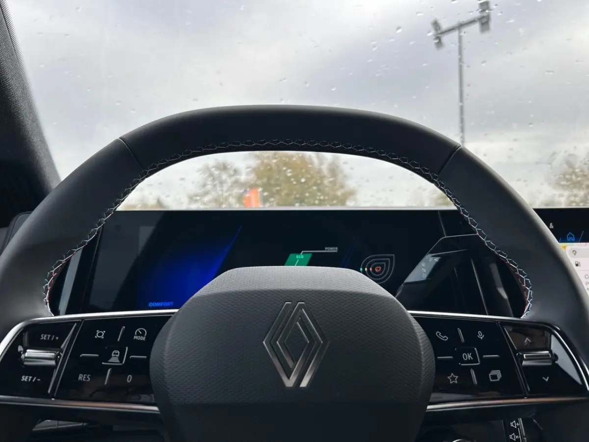 Volant noir avec logo Renault en gros plan, tableau de bord digital lumineux visible derrière sous pluie légère.