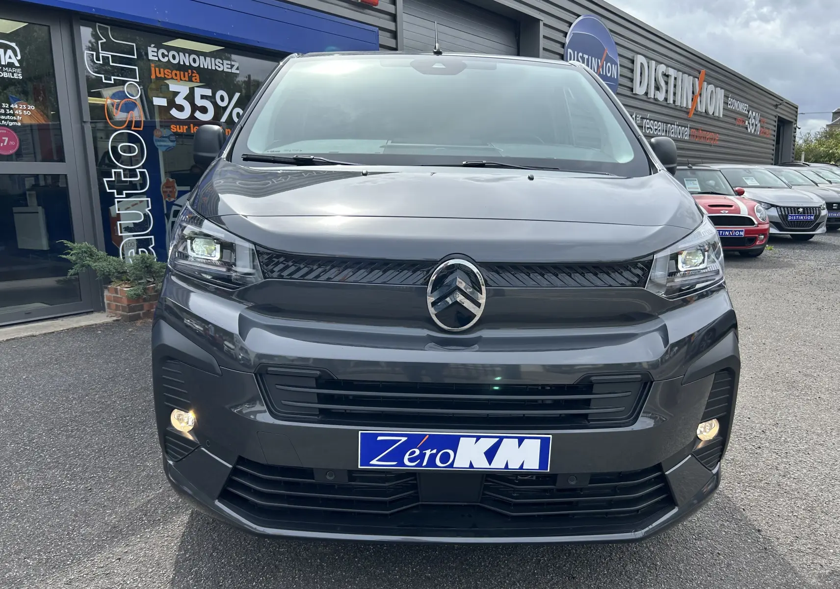 Vue frontale d'un Citroën Jumpy Fourgon gris acier 2025 avec phares allumés et plaque Zéro KM sur un parking commercial.
