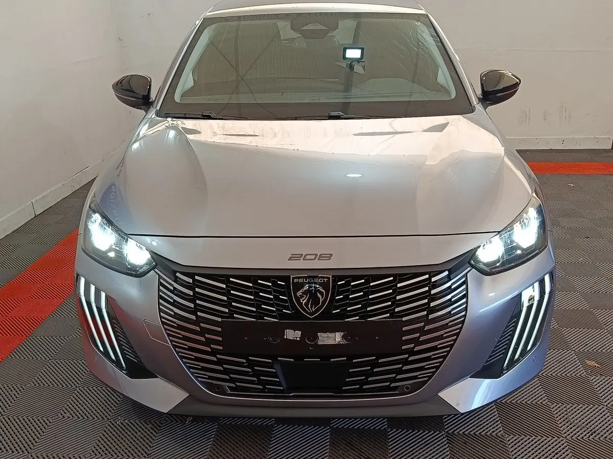 Vue frontale d'une Peugeot 208 gris clair avec phares LED allumés et calandre noire distinctive.
