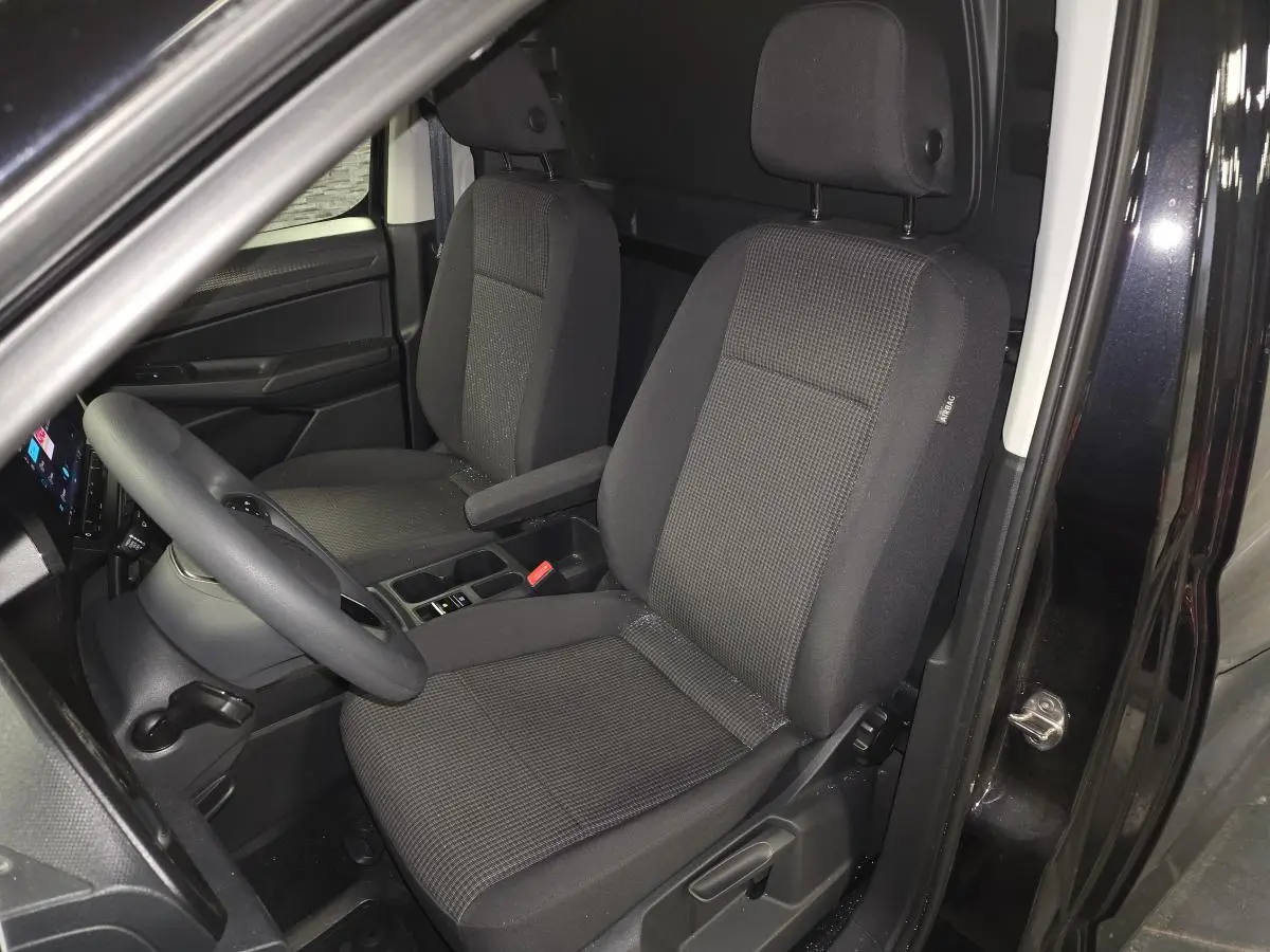 Vue intérieure côté conducteur du Volkswagen Caddy Cargo noir 2024, sièges tissu noir avec accoudoir et tableau de bord visible.