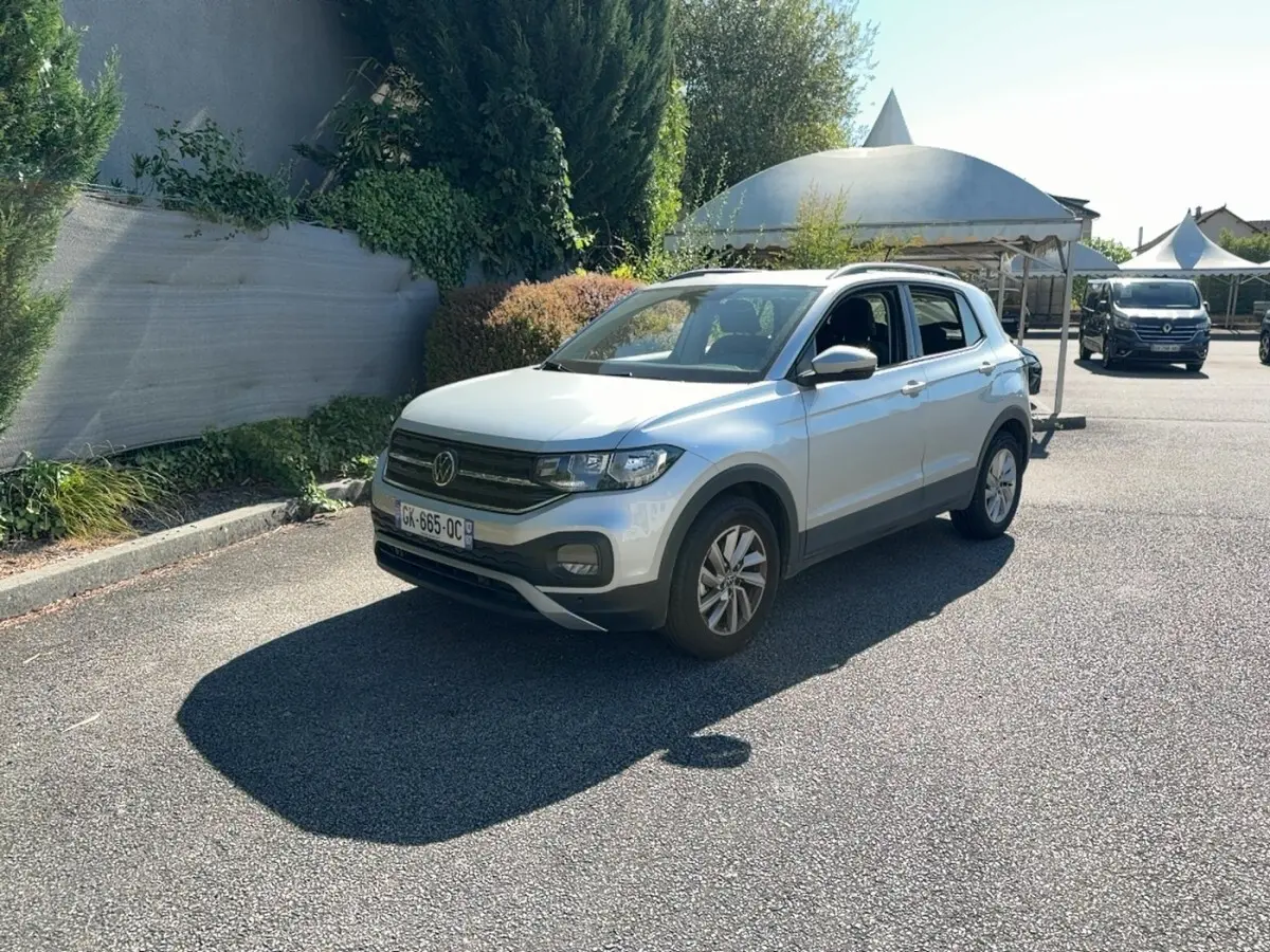 Volkswagen T-Cross gris clair en 3/4 avant droit, stationné sur un parking ensoleillé avec végétation en arrière-plan.