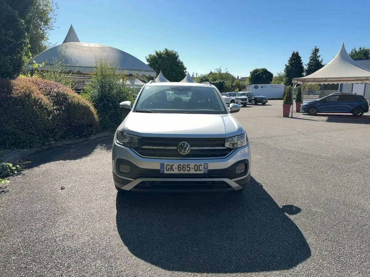 Volkswagen T-Cross gris clair vue de face, avec calandre noire et feux LED, stationné en extérieur sous un ciel bleu.