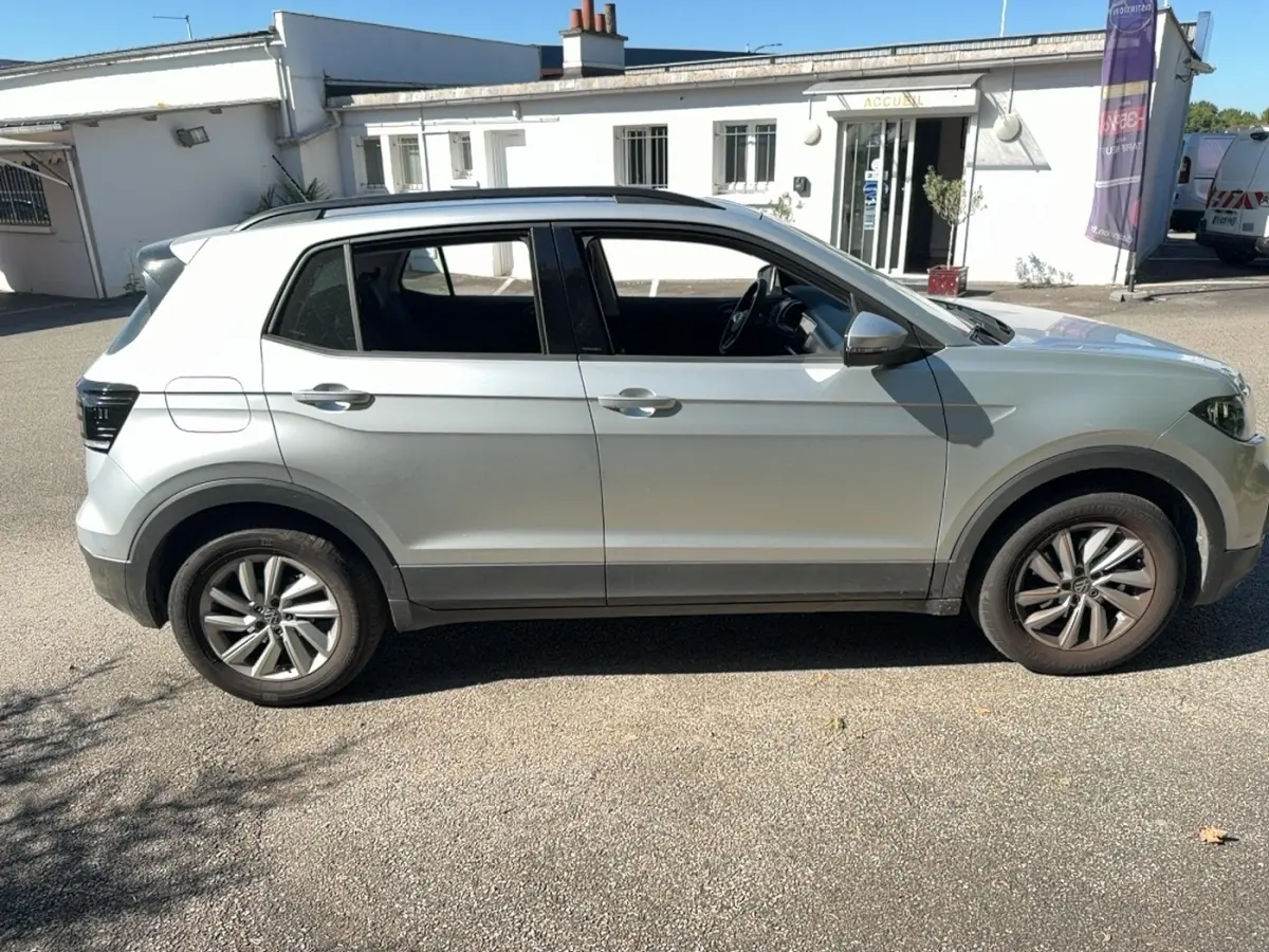 Vue de profil côté gauche d’un Volkswagen T-Cross gris clair avec jantes alliage et barres de toit noires.