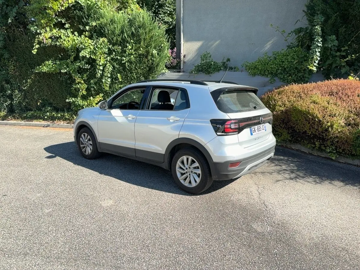 Volkswagen T-Cross gris clair vue 3/4 arrière droit, avec barres de toit noires et jantes alliage.