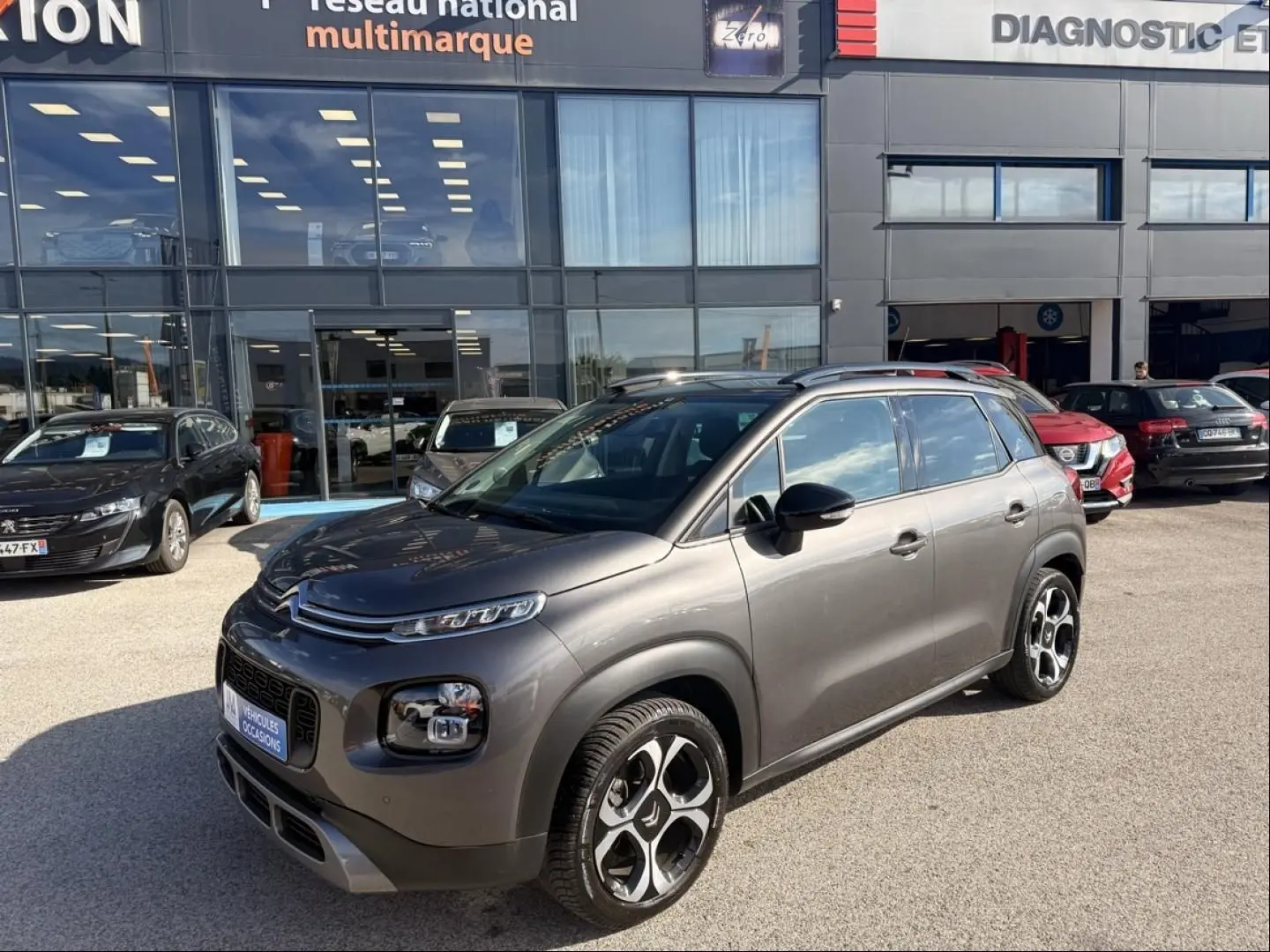 Citroën C3 Aircross gris métal en 3/4 avant droit, avec jantes distinctives et barres de toit visibles.