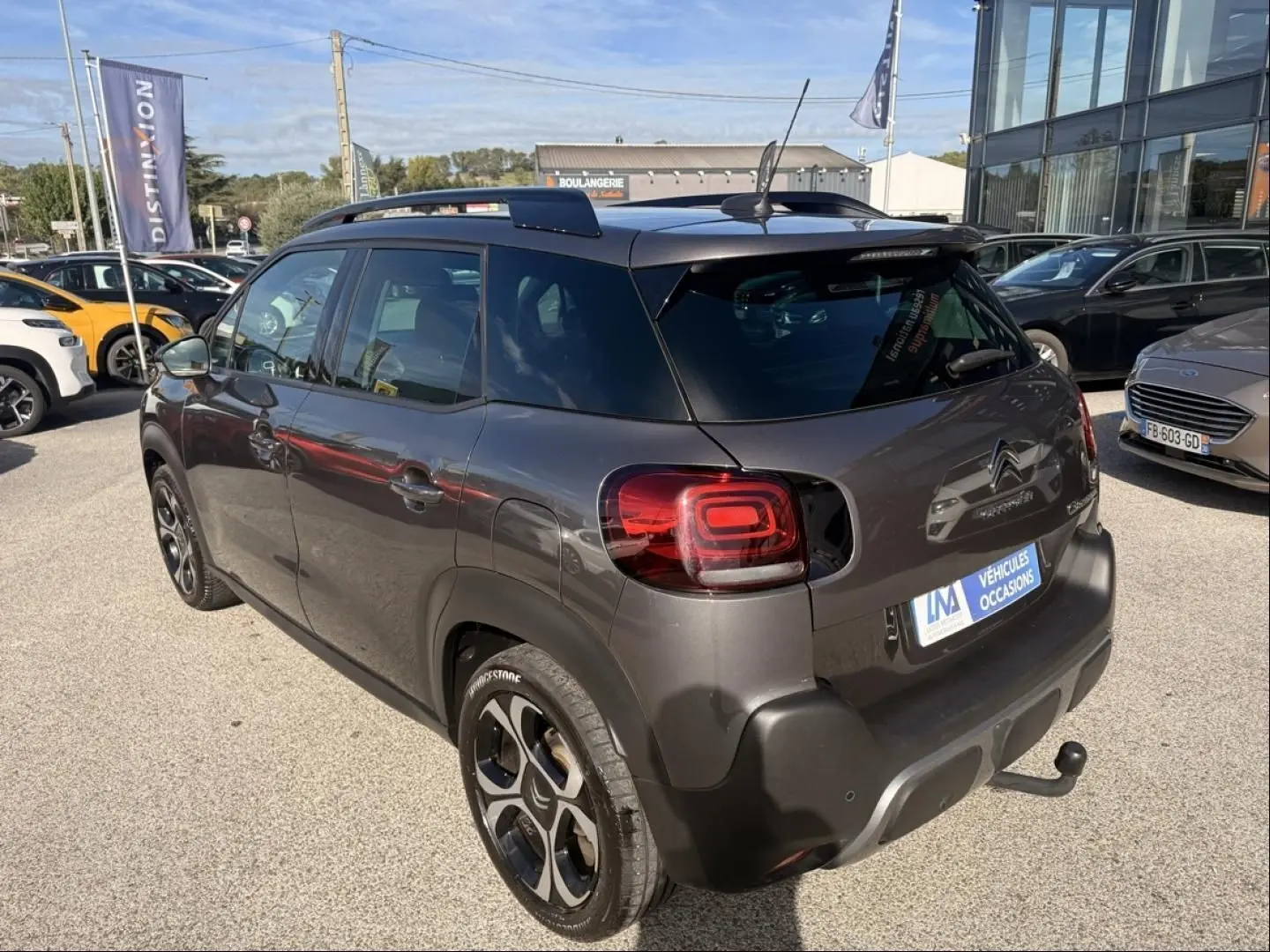Citroën C3 Aircross gris métal vue 3/4 arrière droit, avec barres de toit et attelage visible.