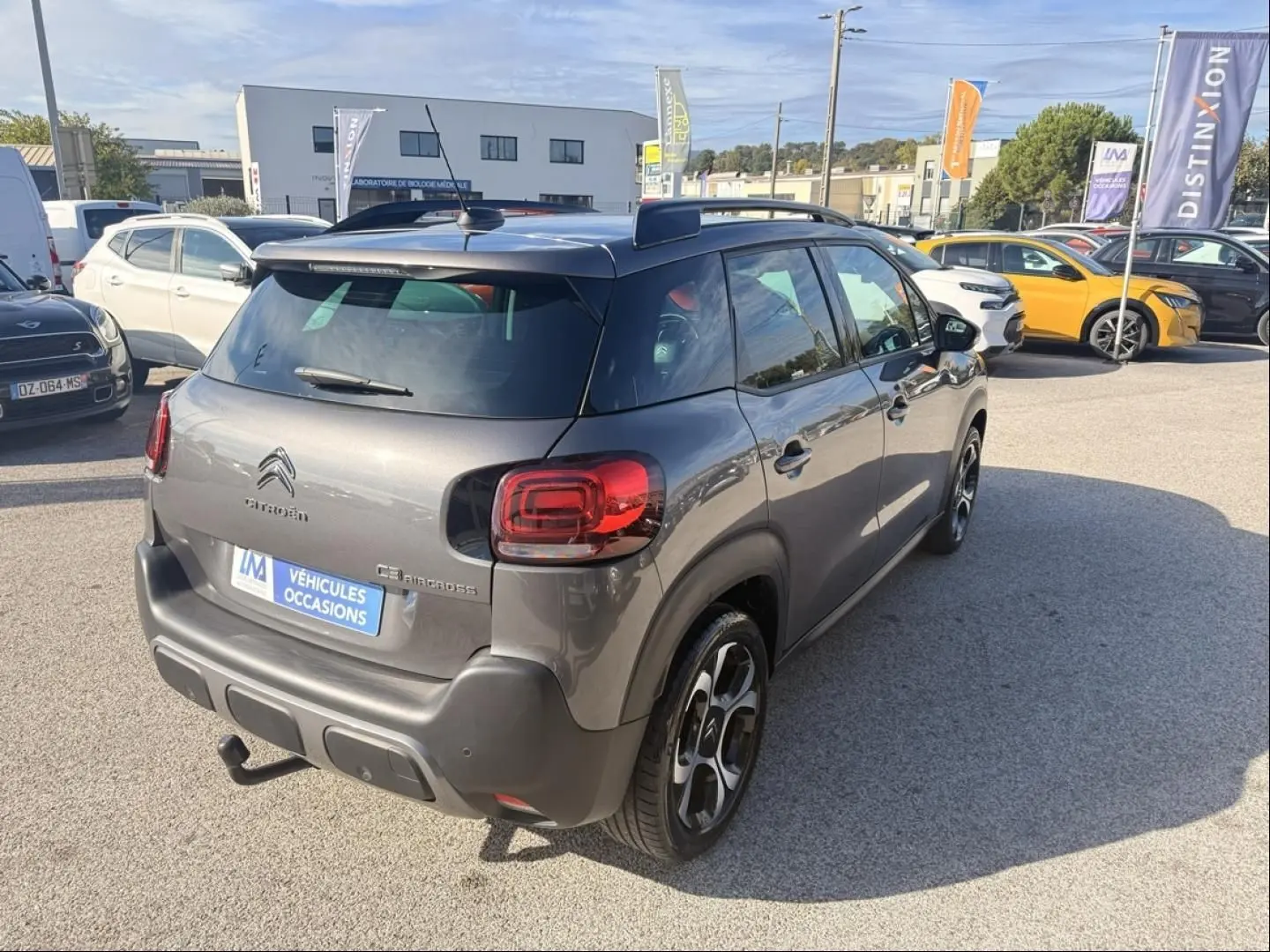 Vue 3/4 arrière droite d'un Citroën C3 Aircross gris métal 2021 avec barres de toit et attelage visible sur parking.