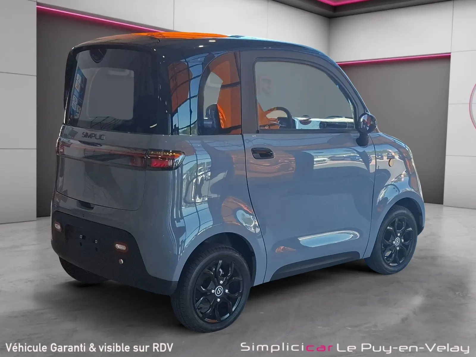 Vue 3/4 arrière droite d'une Simplici S1 électrique gris perle avec toit noir et jantes noires en intérieur showroom.