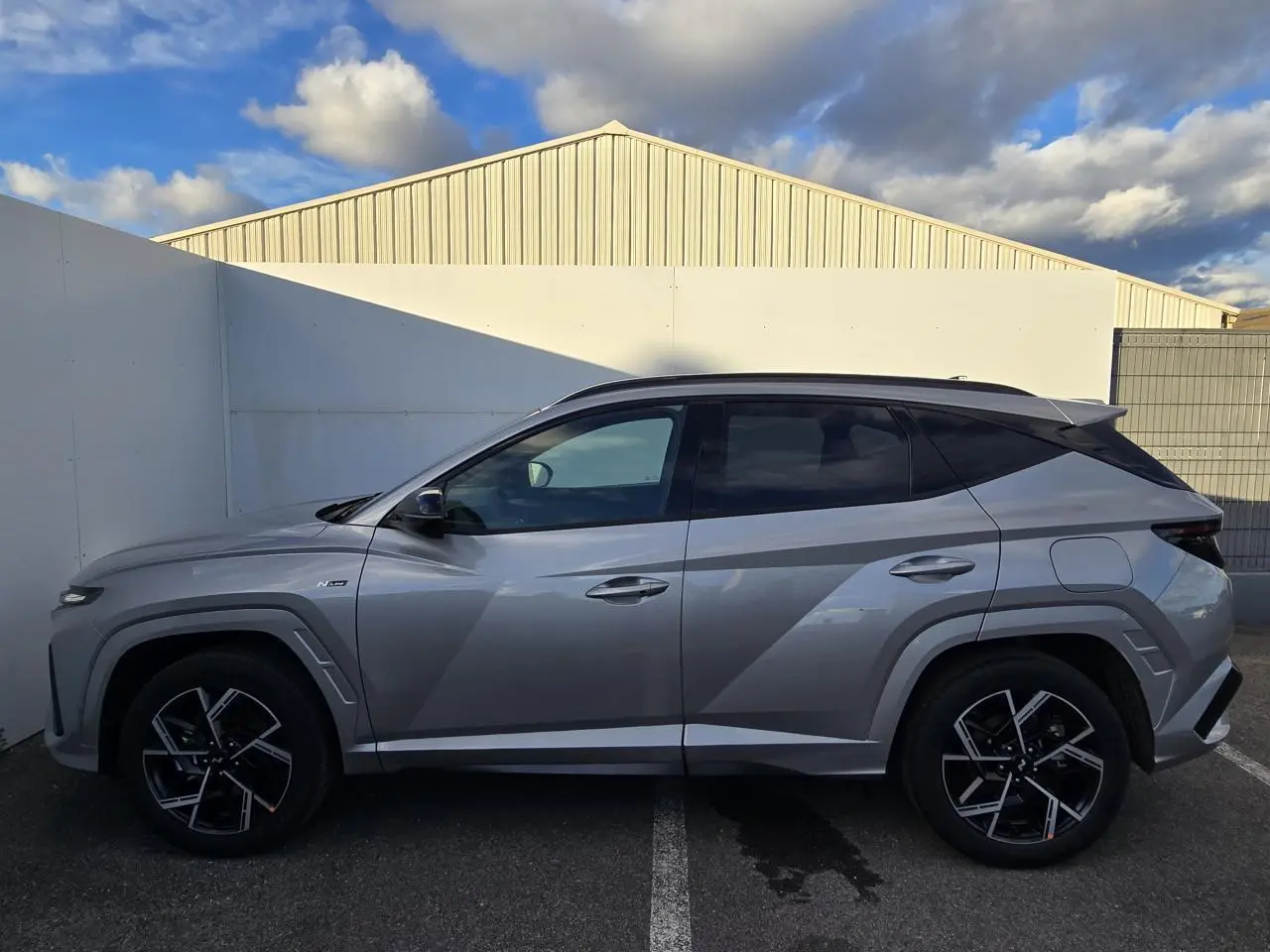 Vue de profil côté gauche d'un Hyundai Tucson 2025 gris métallisé avec jantes alliage noires et toit noir.