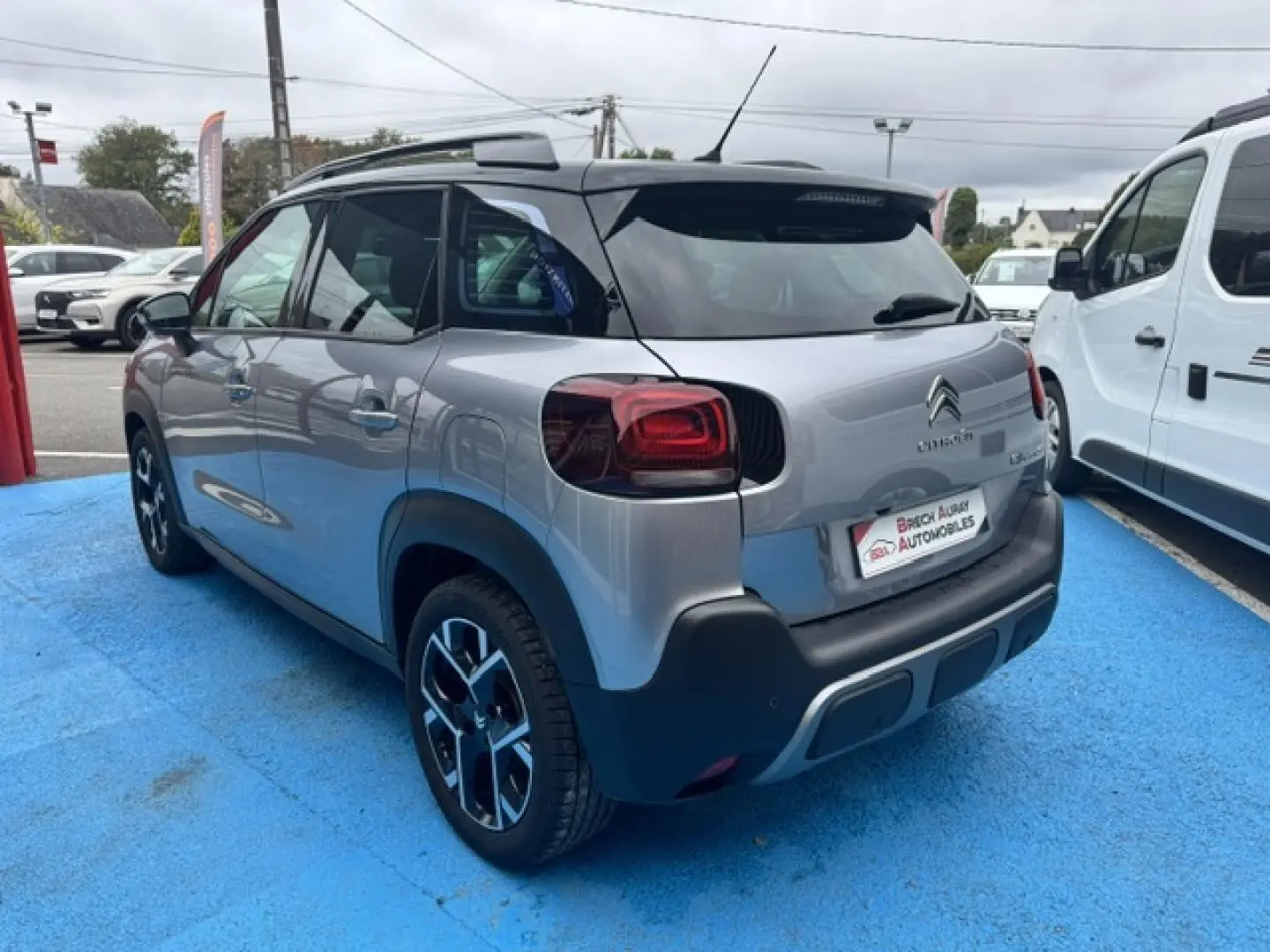 Vue 3/4 arrière droite d'une Citroën C3 Aircross gris acier avec barres de toit et jantes alu noires.