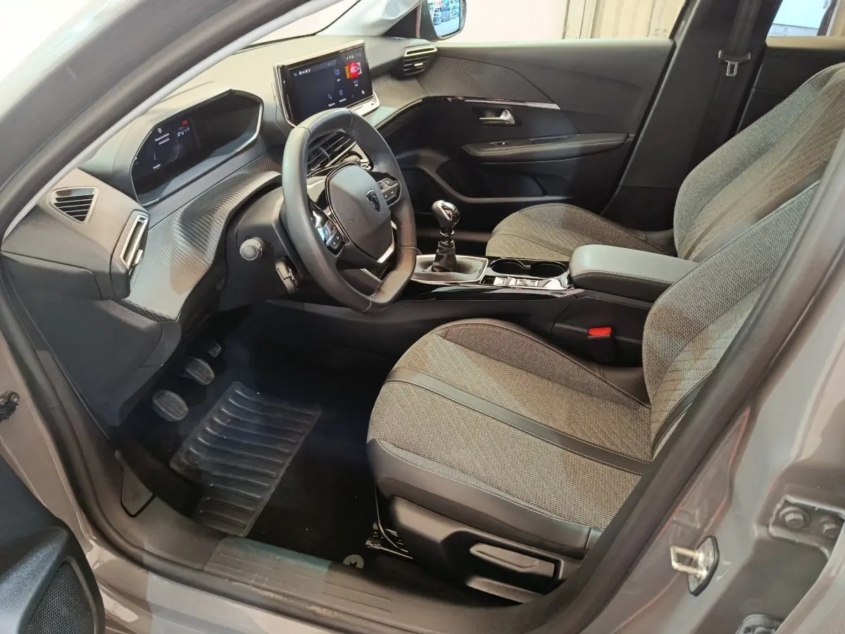 Intérieur de la Peugeot 208 PureTech 100 S&S Allure 2025 en gris foncé, vue côté conducteur avec tableau de bord et sièges tissu.