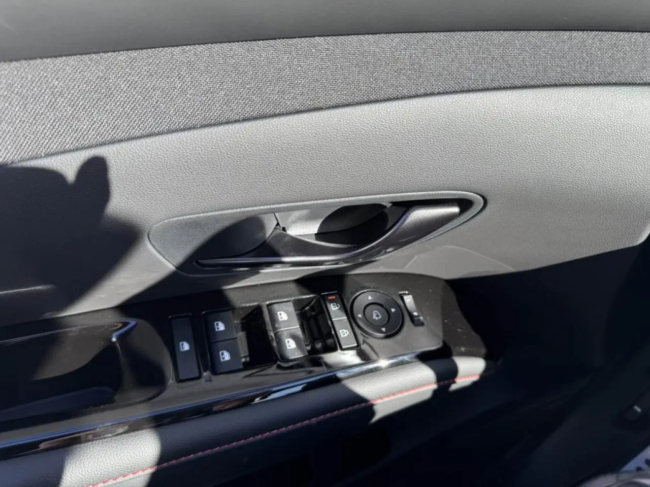 Gros plan sur la commande de lève-vitres et poignée intérieure côté conducteur du Hyundai Tucson noir avec surpiqûres rouges.