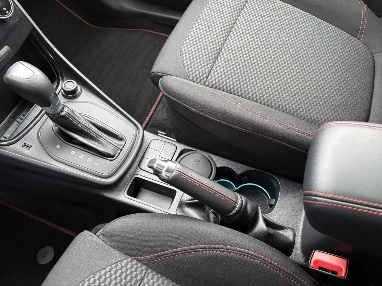 Vue intérieure rapprochée du levier de vitesses automatique et frein à main avec surpiqûres rouges sur sièges noirs.