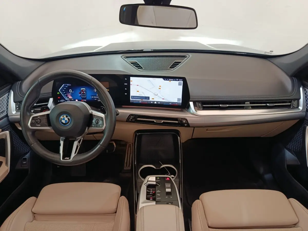 Vue intérieure avant du BMW X1 xDrive25e M Sport 2024 avec tableau de bord beige et écran tactile central lumineux.