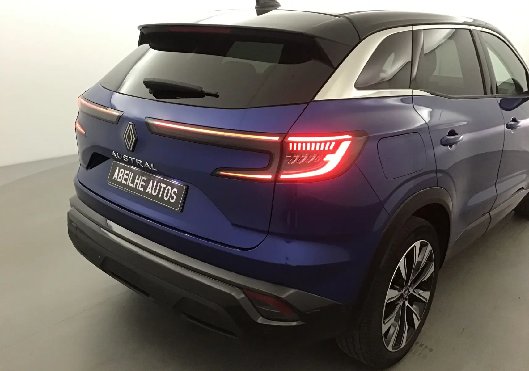 Vue 3/4 arrière droite du Renault Austral bleu Iron avec toit noir et feux arrière allumés sur fond neutre.