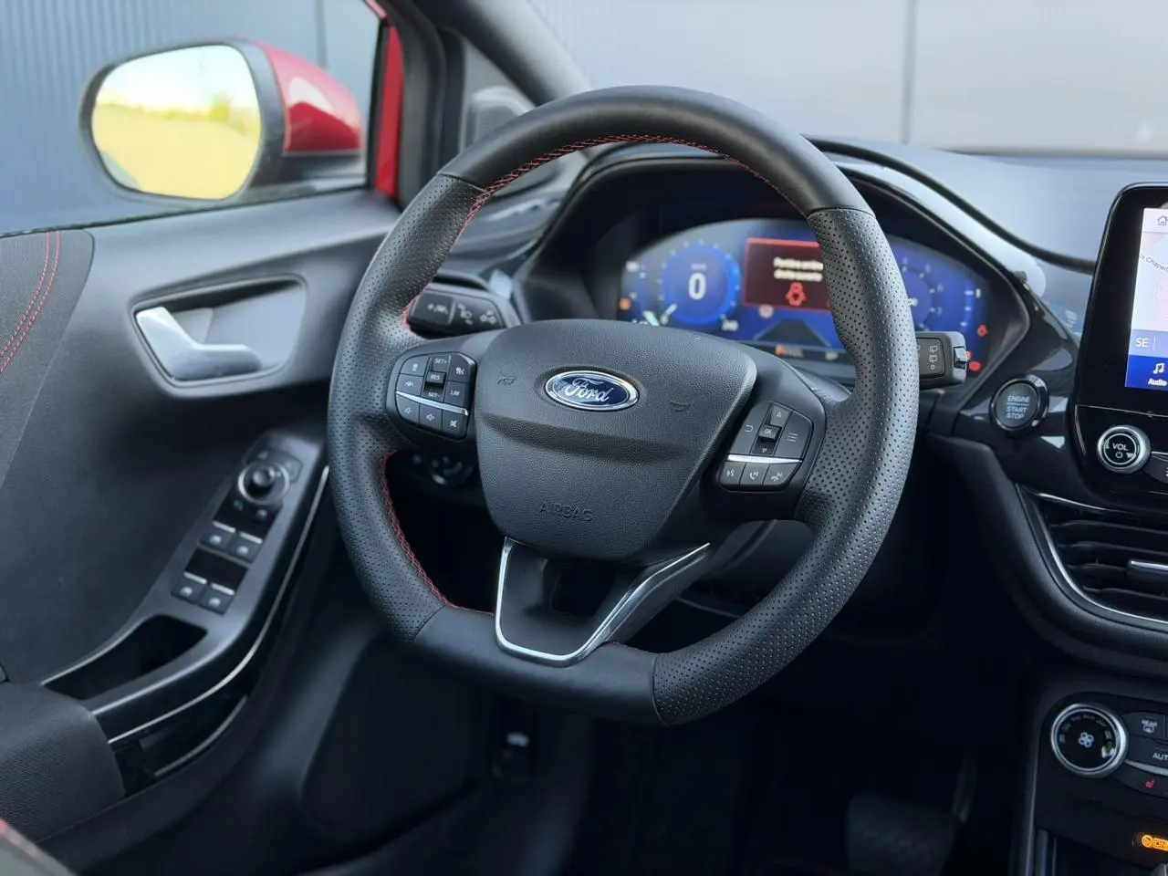 Vue intérieure centrée sur le volant ST-Line noir avec surpiqûres rouges, tableau de bord numérique et commandes sur porte côté gauche.