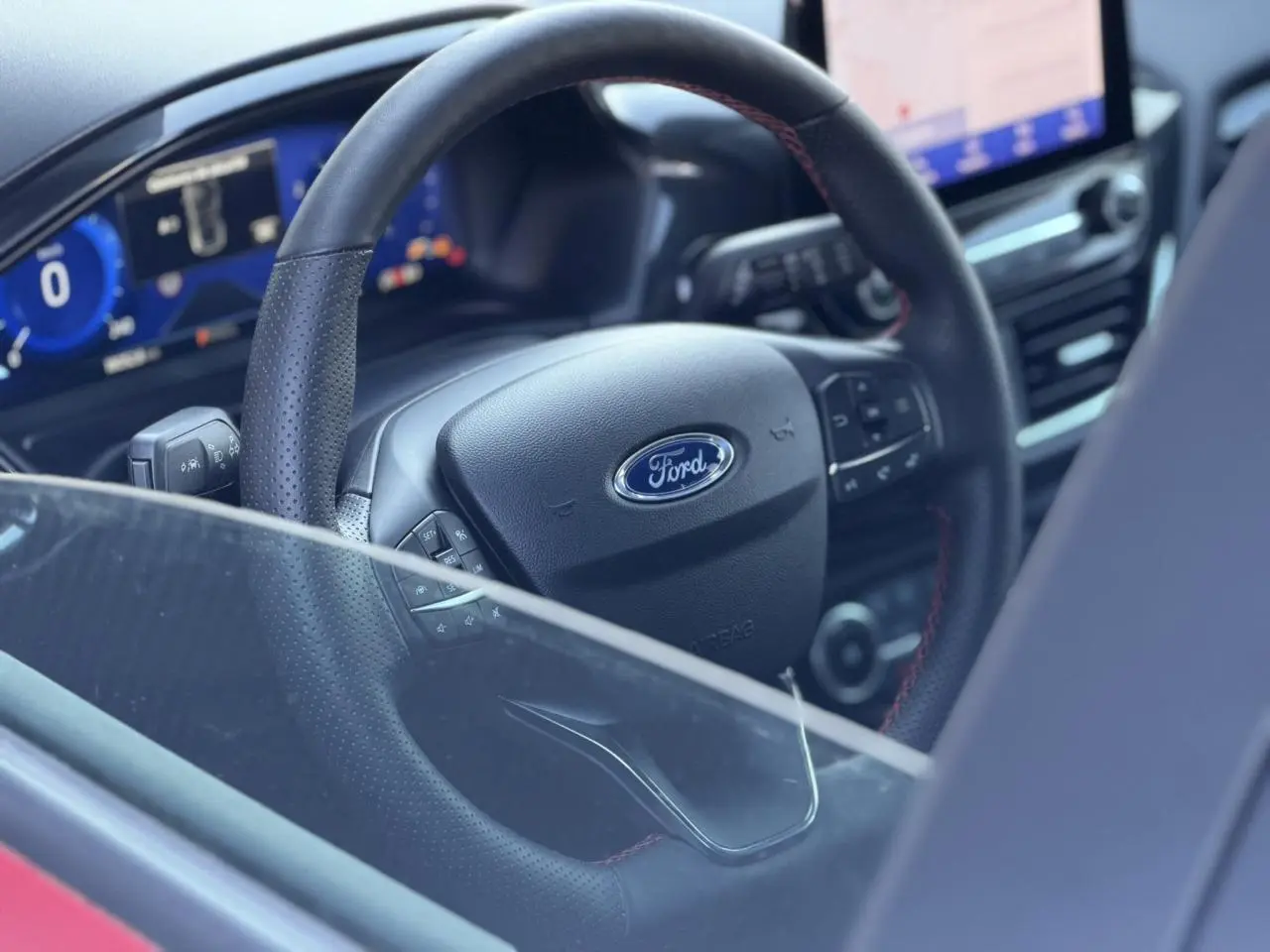 Vue rapprochée de l’intérieur du Ford Puma 2023, volant ST-Line en cuir noir avec surpiqûres rouges visible à travers la vitre partiellement baissée.