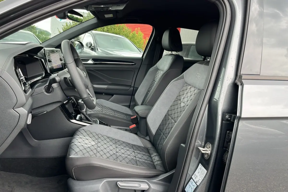 Vue intérieure côté conducteur du Volkswagen T-Roc gris indium 2025, sièges tissu R-Line avec surpiqûres et volant cuir multifonctions.
