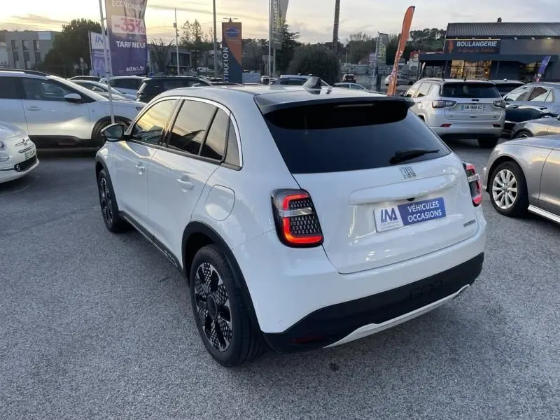 Vue 3/4 arrière droite d'une FIAT 600 blanche 2025 hybride, avec feux arrière allumés et jantes noires distinctives.