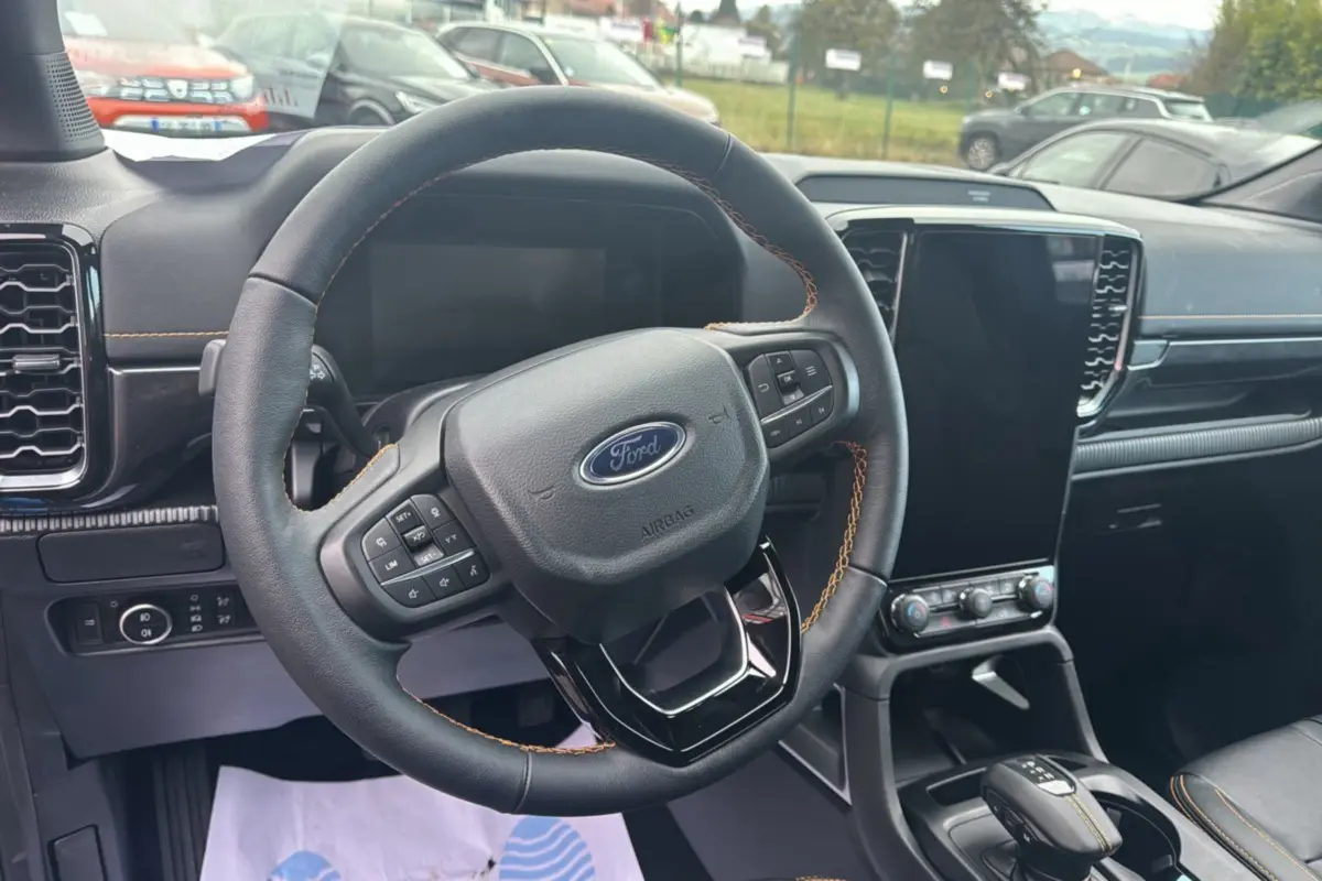 Vue intérieure centrée sur le volant cuir noir avec surpiqûres orange du Ford Ranger 2025, écran tactile vertical et console centrale.