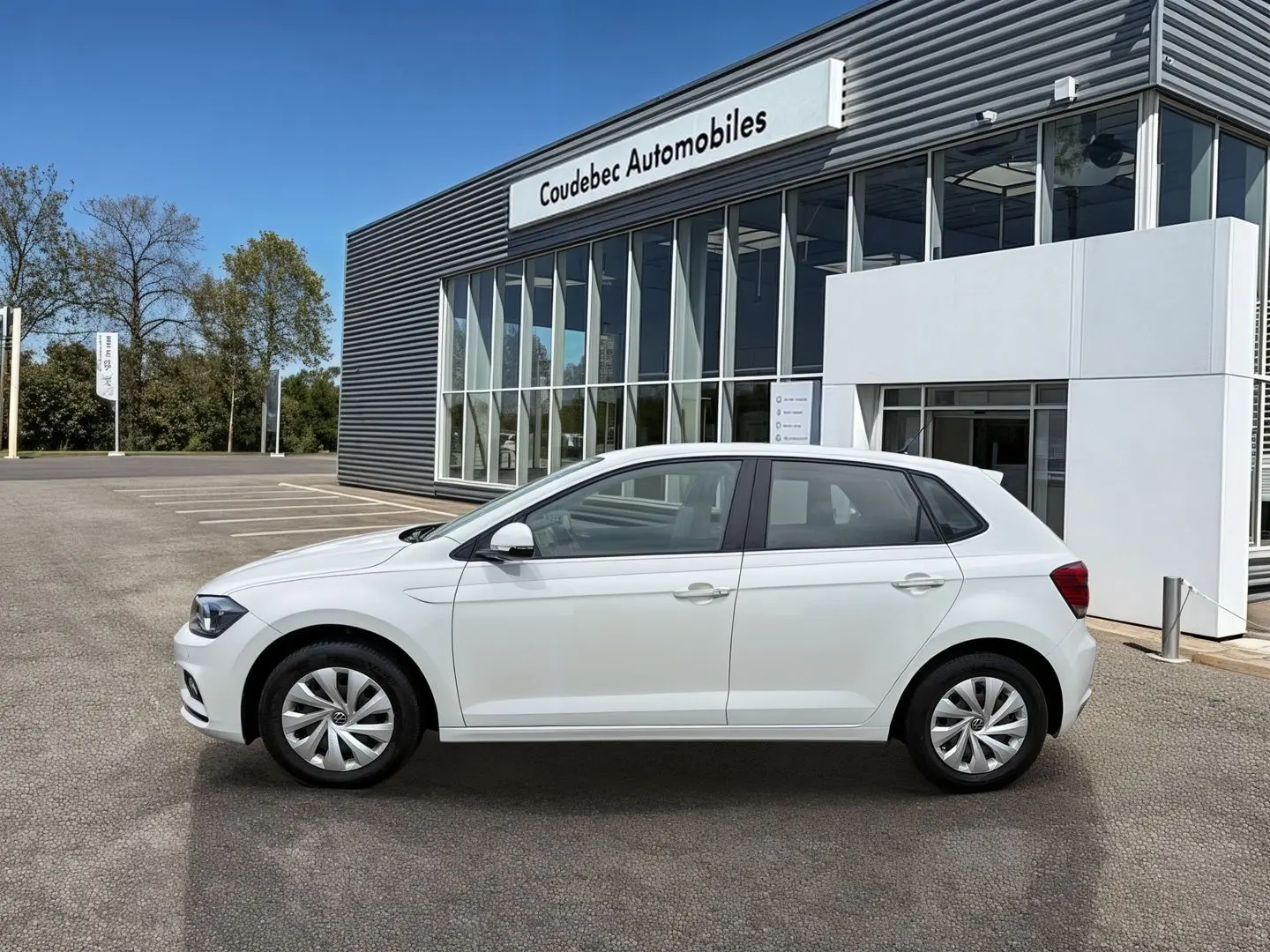 Vue latérale droite d'une Volkswagen Polo Business blanche stationnée devant un bâtiment moderne avec jantes en acier et enjoliveurs intégrals.