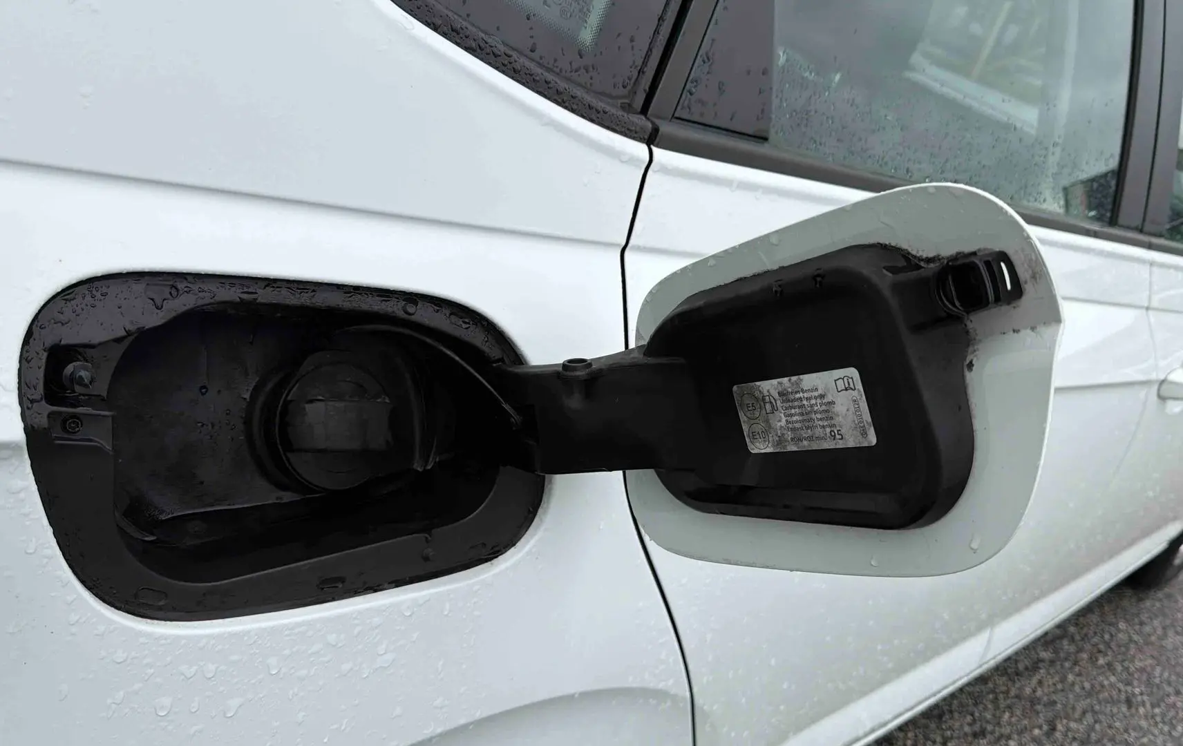 Gros plan sur la trappe à carburant ouverte côté gauche d’une Volkswagen Polo Business blanche sous la pluie.
