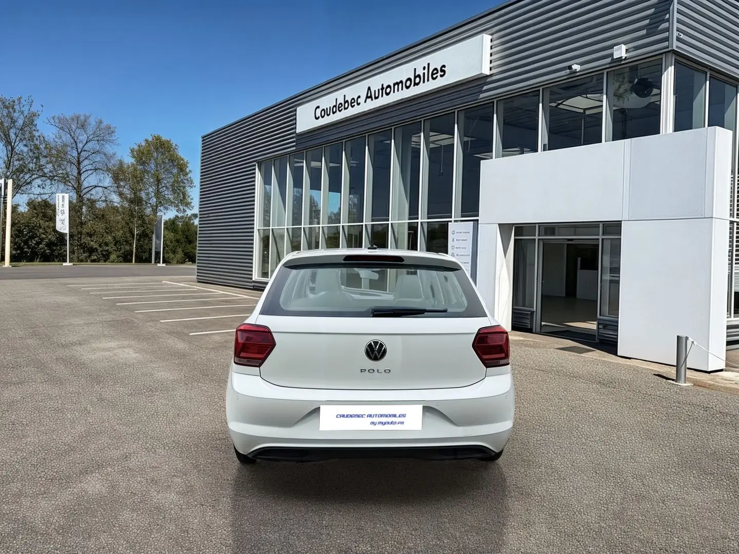 Vue arrière d'une Volkswagen Polo Business blanche stationnée devant un bâtiment commercial moderne.