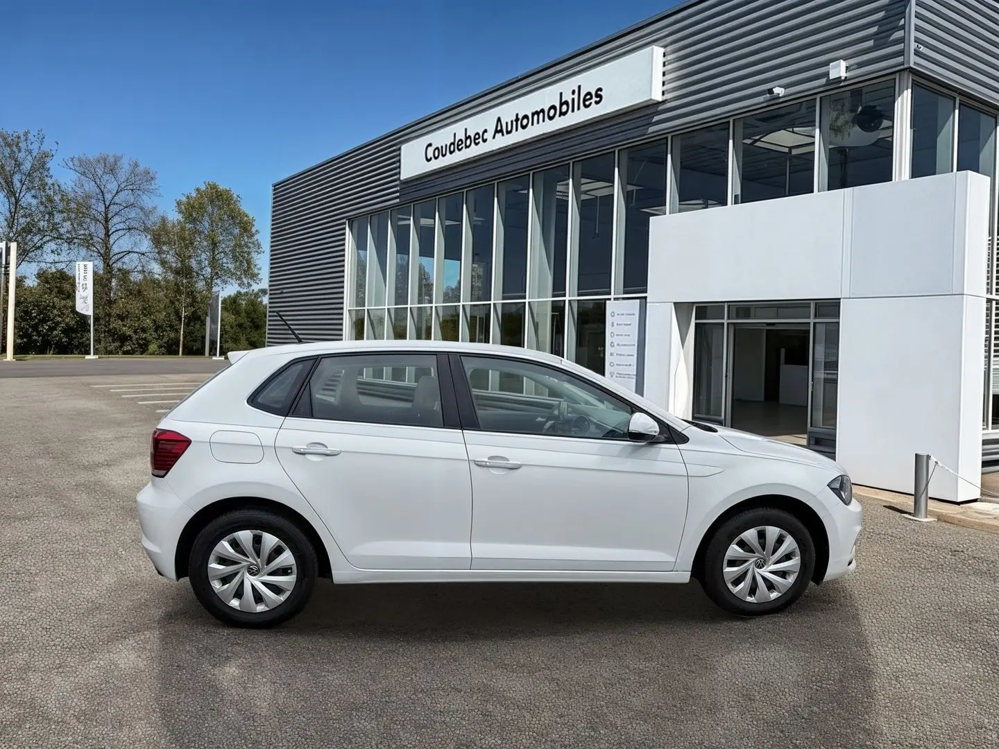 Vue de profil côté gauche d'une Volkswagen Polo Business blanche stationnée devant un bâtiment commercial.