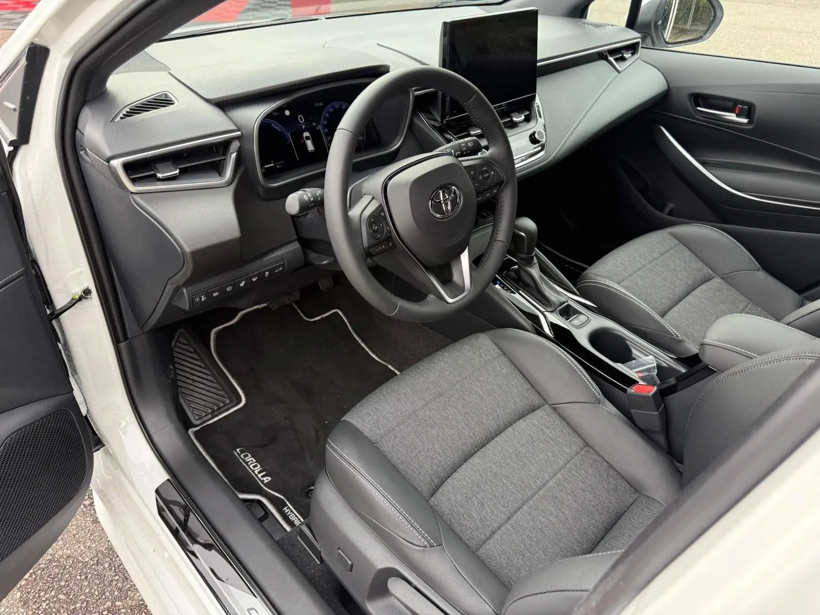 Vue intérieure côté conducteur de la Toyota Corolla Touring Sport blanche, avec tableau de bord moderne et sièges gris tissu et cuir.