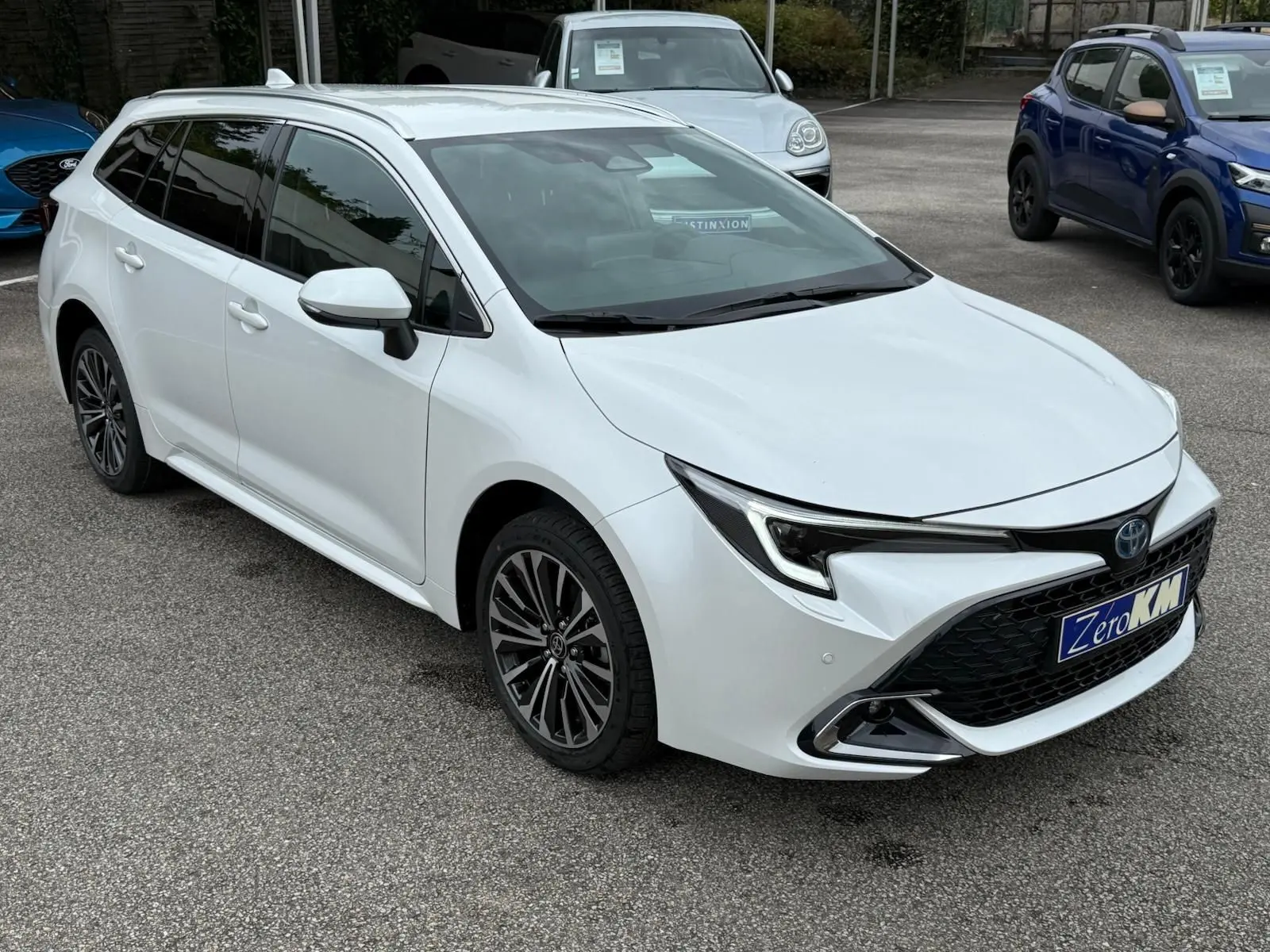 Toyota Corolla Touring Sport blanche nacrée vue en 3/4 avant droit sur parking avec jantes alliage et calandre noire.