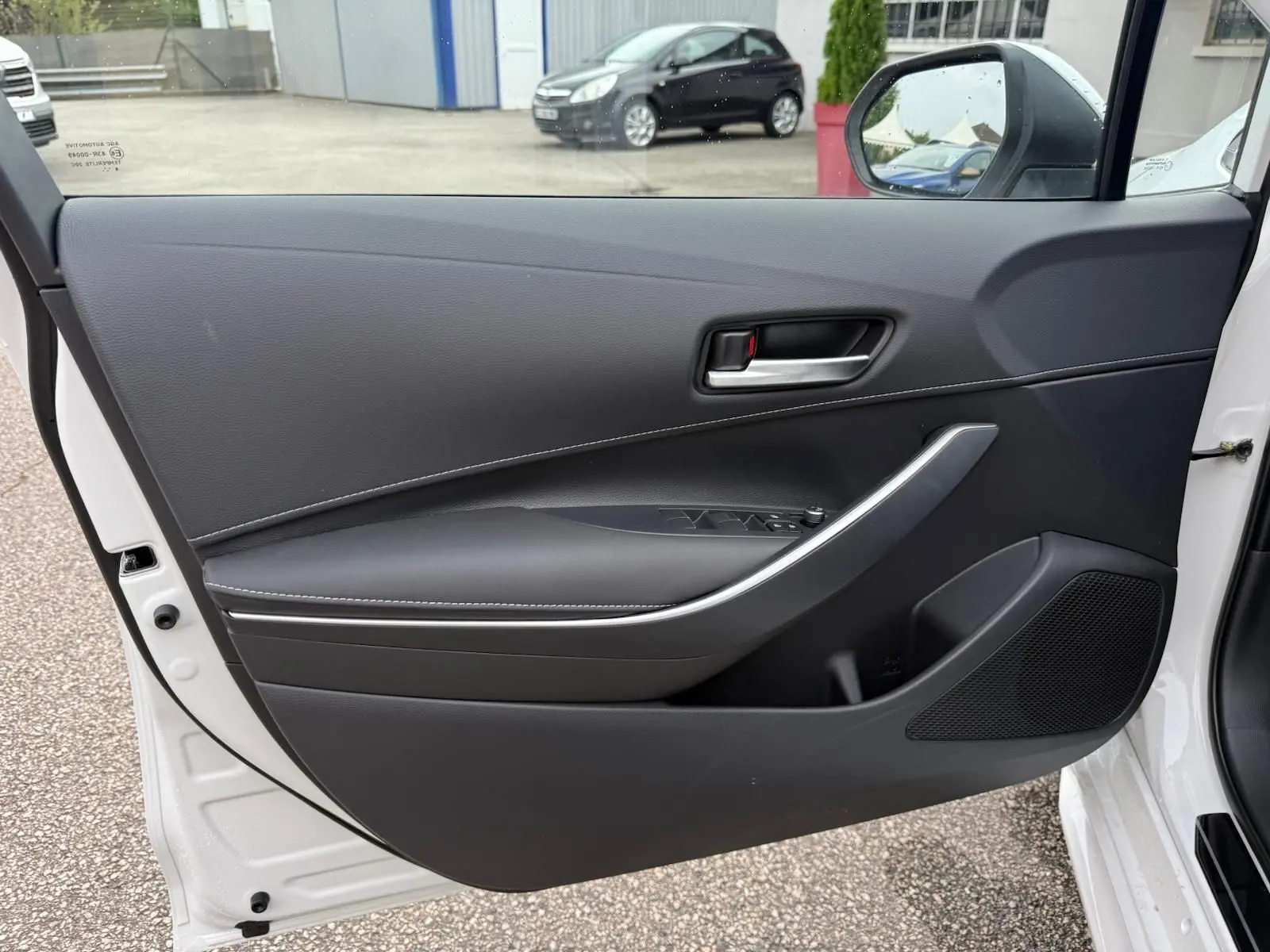 Porte avant gauche ouverte d'une Toyota Corolla Touring Sport blanche, intérieur noir avec poignée argentée et commandes de vitres.