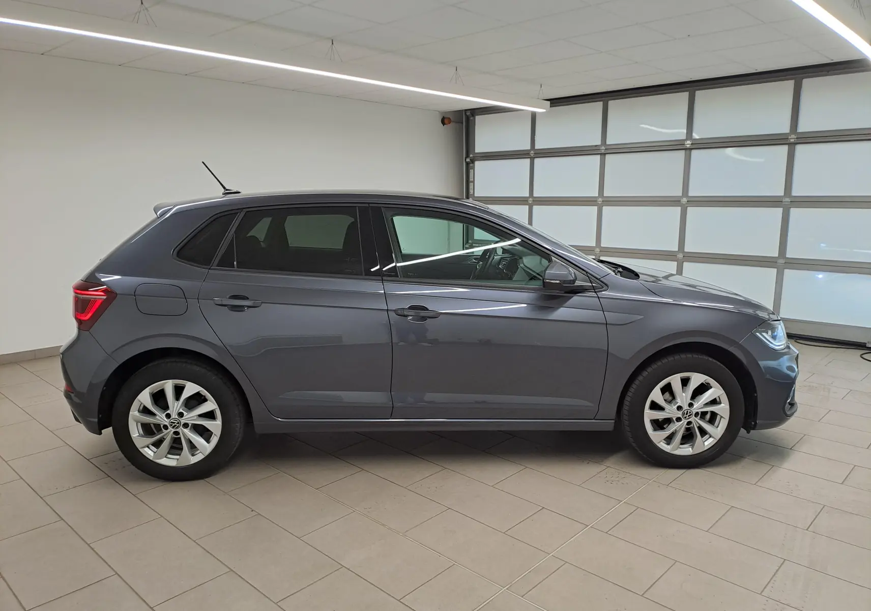 Profil droit d'une Volkswagen Polo 2023 gris cendré avec vitres arrière surteintées dans un garage lumineux.