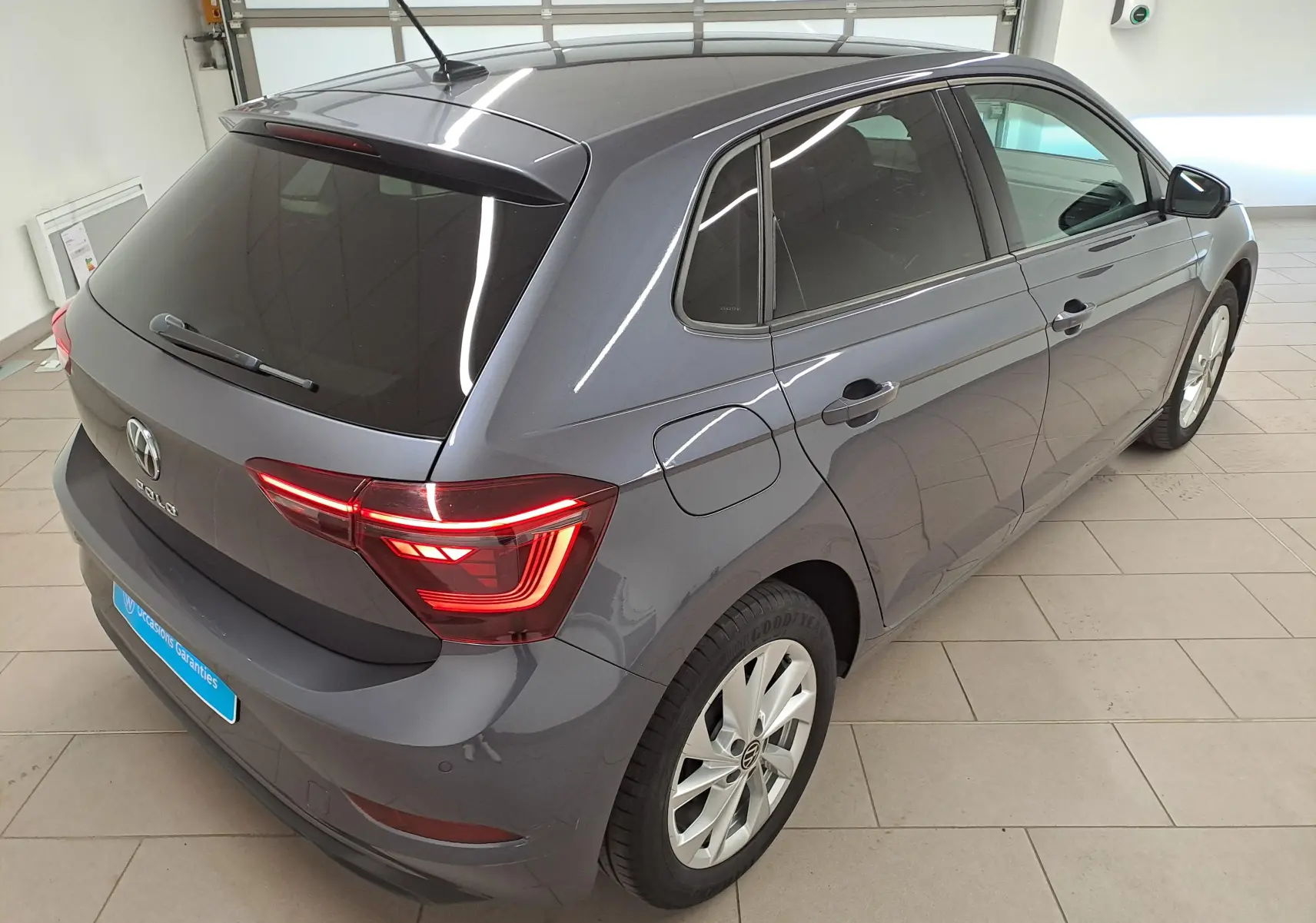 Vue 3/4 arrière droite d'une Volkswagen Polo gris cendré 2023 avec vitres arrière surteintées et feux arrière allumés.