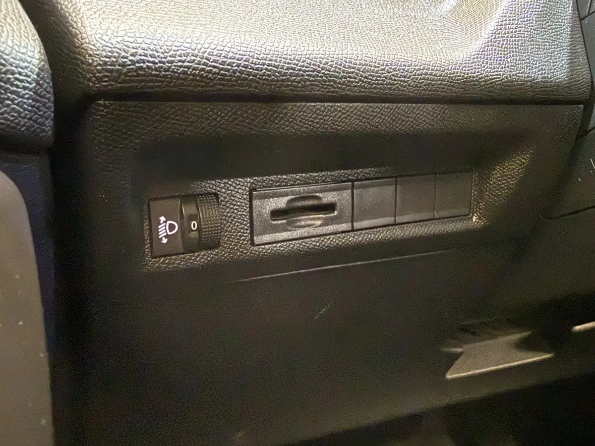 Détail intérieur montrant les commandes de réglage des phares sur le côté gauche du tableau de bord d’une Peugeot 308 gris foncé.