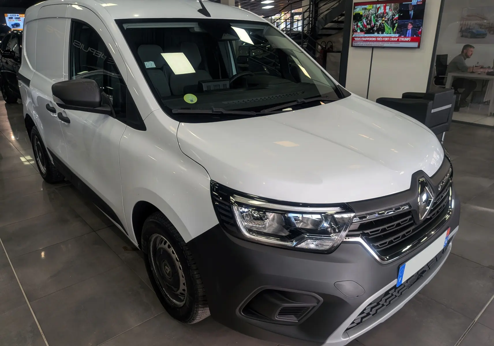Vue 3/4 avant d'un Renault Kangoo Van blanc minéral avec pare-chocs noir mat et jantes acier en intérieur showroom.