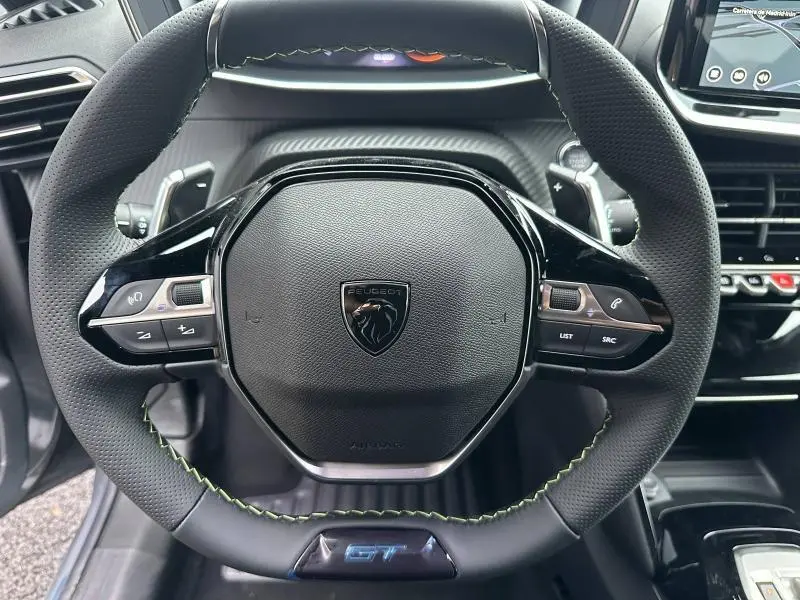 Vue rapprochée du volant noir perforé avec surpiqûres vertes et logo Peugeot sur le modèle 208 Hybrid GT 2025.