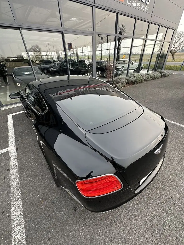 Vue 3/4 arrière droite d'une Bentley Continental GT Mulliner noire brillante, avec feux arrière rouges et logo Bentley visible.