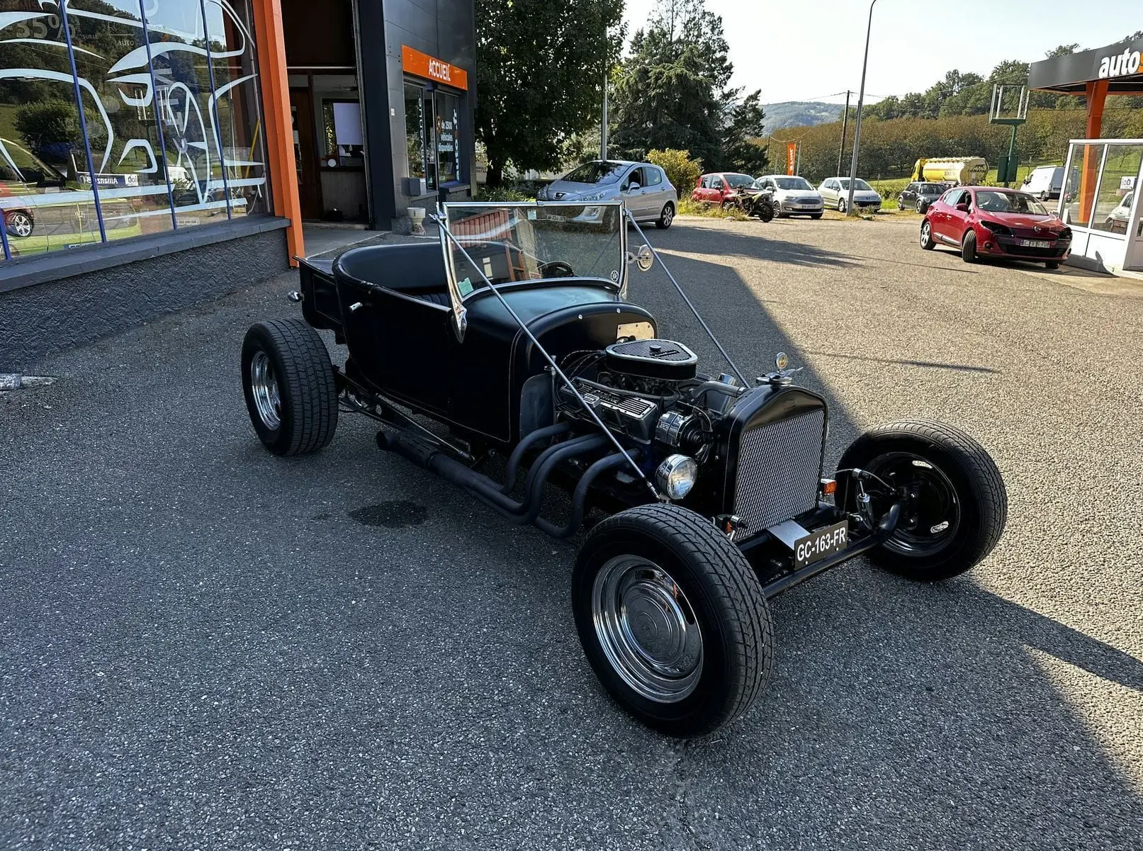 Ford T Hot Rod V8 noir en 3/4 avant droit, capote décapotée et moteur V8 apparent sur parking extérieur.