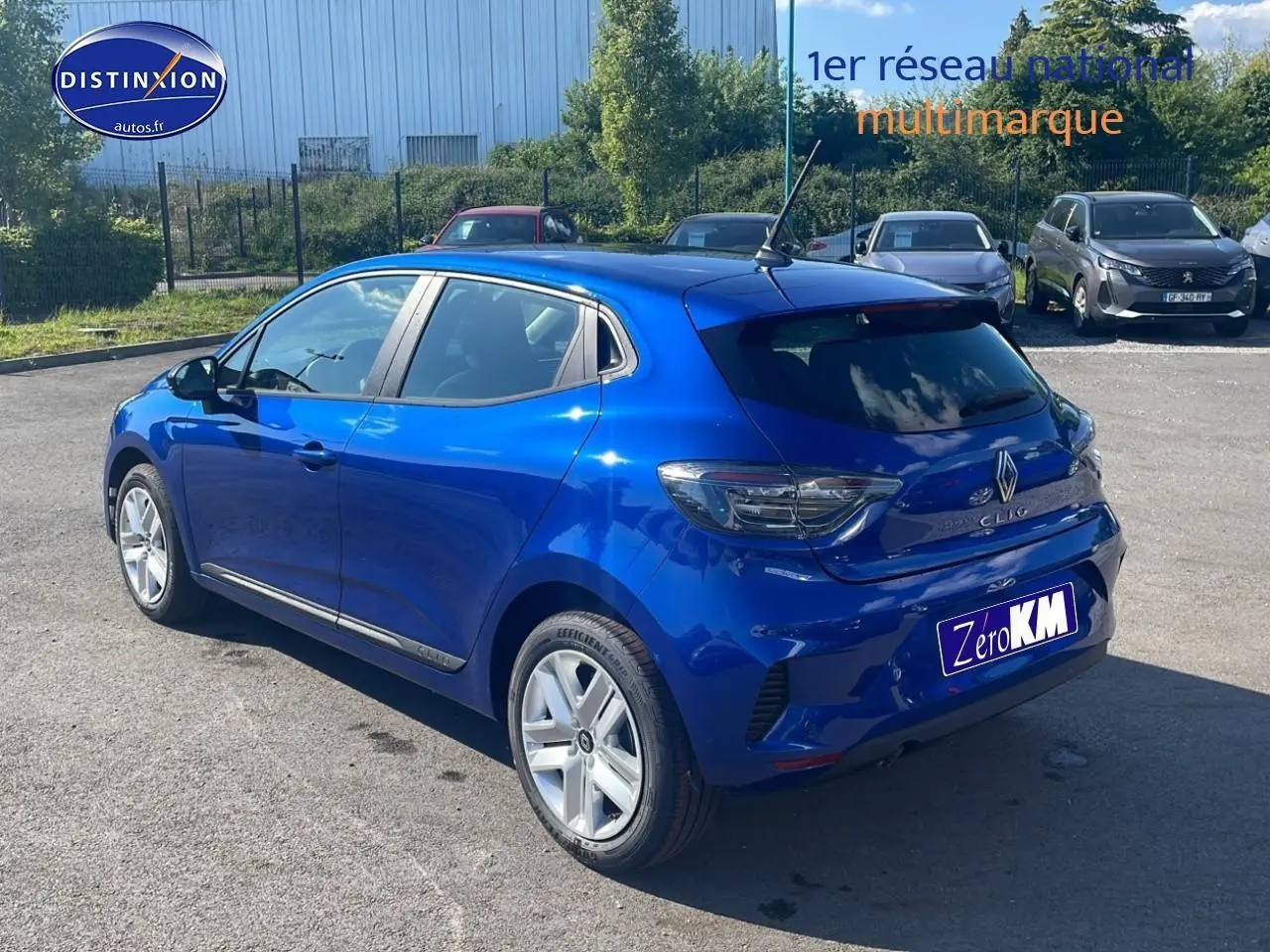 Renault Clio V bleu iron vue 3/4 arrière droit sur parking avec becquet et feux arrière LED teintés.