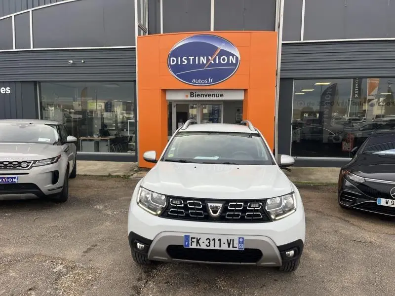 Dacia Duster blanc glacier vu de face, avec calandre noire et barres de toit chromées devant un garage Distinxion.