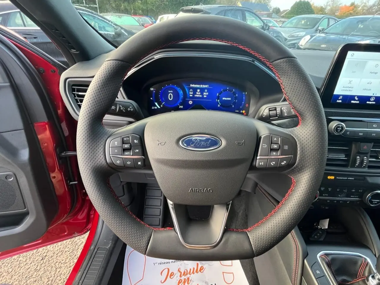 Vue intérieure centrée sur le volant perforé avec surpiqûres rouges du Ford Kuga rouge Lucifer, tableau de bord digital visible.